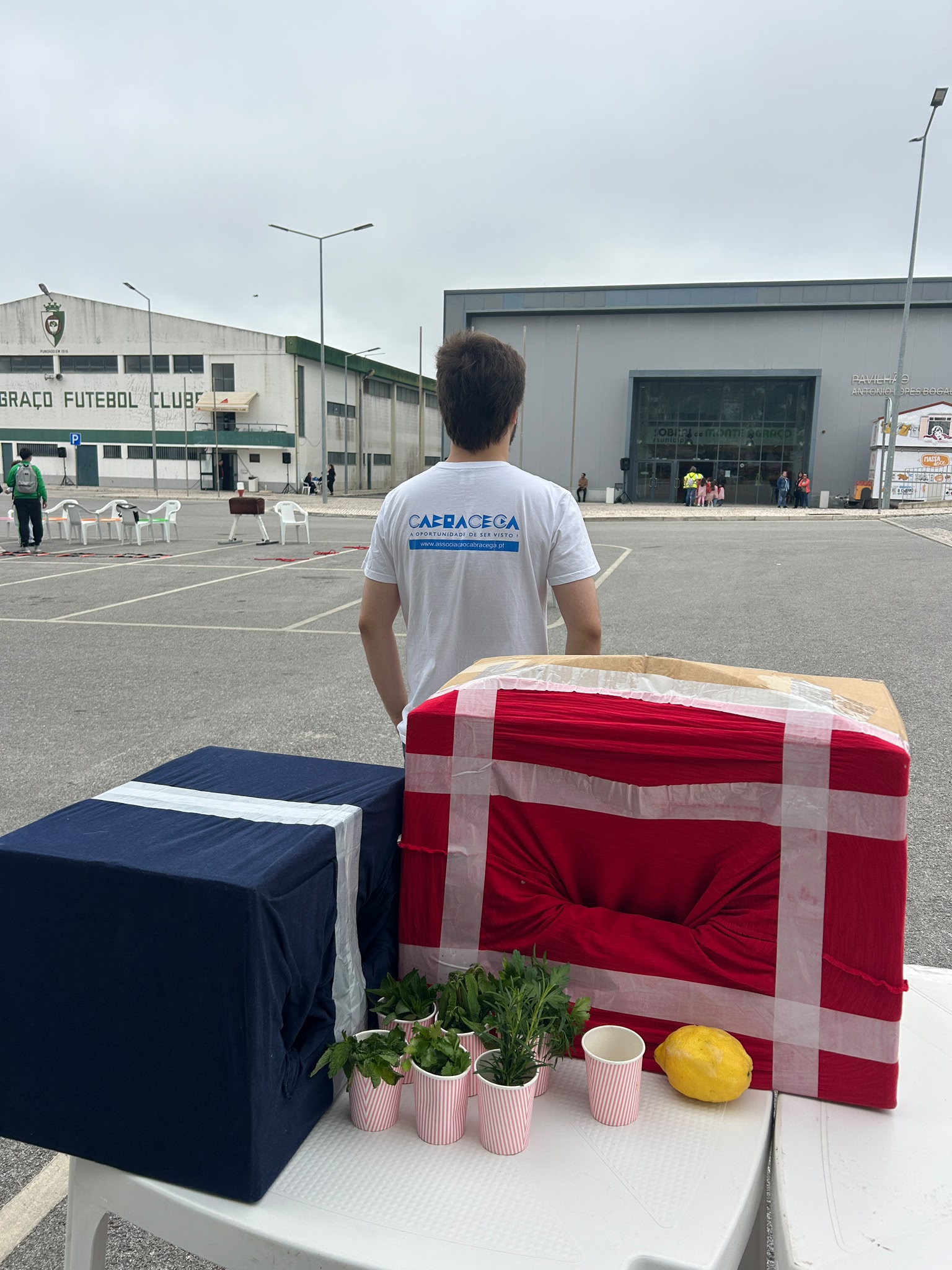 Um jovem de costas vestindo uma T shirt branca está em pé num parque de estacionamento amplo, voltado para dois edifícios grandes à frente. A t shirt tem nas costas o texto “cabracega” em letras grandes azuis e, por baixo, em letras menores, “A oportunidade de ser vista” e o endereço do site da Associação, também em azul, com uma faixa azul horizontal. Em primeiro plano, sobre uma mesa de plástico branca, há duas caixas grandes em forma de cubo: a da esquerda está forrada com tecido azul escuro com uma faixa de fita adesiva branca a cruzar por cima; a da direita é uma caixa de cartão coberta por tecido vermelho com um buraco circular ao centro, franzido, e muitas tiras de fita adesiva transparente formando uma moldura em cruz. À frente das caixas estão vários copos de papel brancos com riscas verticais cor de rosa, cada um com uma pequena planta verde dentro e, ao lado, um limão grande de cor amarelo vivo pousado na mesa. Ao fundo, do lado esquerdo, fica um edifício branco envelhecido com o letreiro “futebol clube” parcial visível, e do lado direito um pavilhão moderno cinzento com fachada de vidro onde algumas pessoas estão agrupadas perto da entrada. Pelo parque de estacionamento estão dispersas algumas cadeiras de plástico e poucas pessoas, e o céu está nublado.